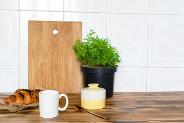 white espresso coffee cup, fresh baked croissant on kitchen table, counter, utensils dishware, home green plant in pot on wooden shelf. Early morning french home breakfast