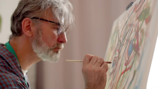 Male artist in glasses with gray hair and beard paints on canvas with brush in workshop, side view. Man paints on canvas at workshop