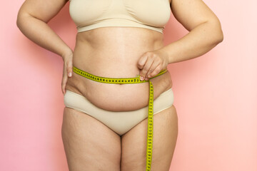 Cropped photo of fat woman tummy in underwear trying to lose weight. Measuring by roulette tape belly, waistline, abdomen. Excess folds on her stomach. Health care, obesity and overweight. Close-up. 