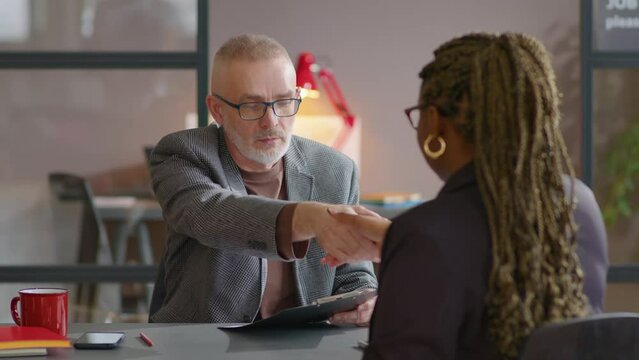 Senior recruiter holding CV, talking and giving handshake to African American female candidate during job interview in office