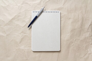Gray notepad with white coiled spring and pen on a background of beige crumpled craft paper. With...