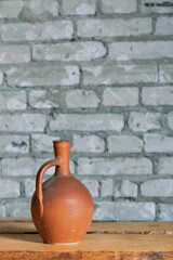 A clay ceramic jug with a high narrow neck against a gray brick wall. The jug stands on a table made of a wide rough board