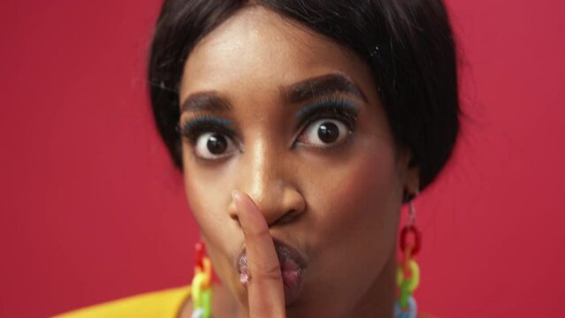 Secret hush. Keep quiet. Stop talking. Shocked surprised scared woman with colorful face makeup warning with shh finger gesture isolated on red.