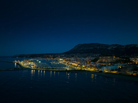 Denia on the Costa Blanca in the evening after the blue hour. A drone shot from the sea overlooking the harbour.