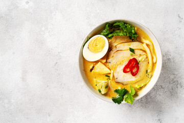 Asian laksa soup with chicken meat in a ceramic bowl on a culinary background. Spicy soup with coconut milk broth, rice noodles and egg in a plate on the kitchen table	