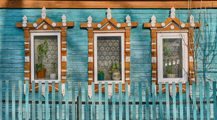 Windows on turquoise plank wall of old house with wooden carved architraves with cracked paint. - obrazy, fototapety, plakaty