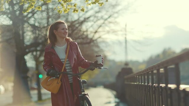 happy elegant woman in red rain coat with bicycle and cup of coffee walking outside in the city.