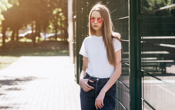 Stylish hipster girl wearing white t-shirt and pink glasses posing against street , urban clothing style.