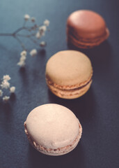 A dark, moody image of chocolate, caramel, and vanilla macaroons arranged on a slate tile, atop a dark grey concrete textured surface background.