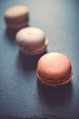A dark, moody image of chocolate, caramel, and vanilla macaroons arranged on a slate tile, atop a dark grey concrete textured surface background.