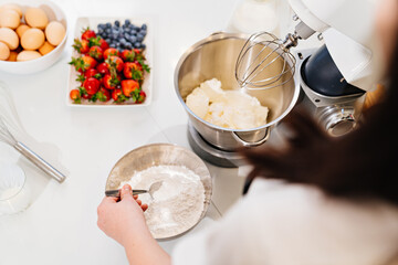 top view. A cook adds a spoonful of flour to prepare dough in a mixer bowl.
