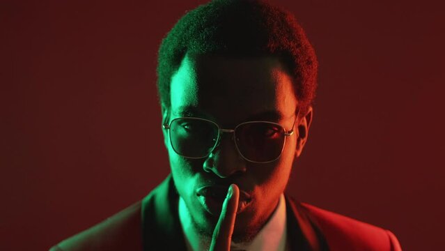 Stop talking. Gentleman secret. Confidential conspiracy. Neon light confident man in suit glasses warning with hush gesture isolated on dark red.