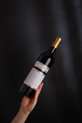 Fototapeta premium hand holding bottle of red wine with mock up label, no brand on black background. Cabernet sauvignon, merlot, pinot noir. Alcohol drink, copy space