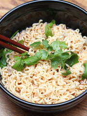 Instant noodles in a black bowl with coriander sprinkles served with chopsticks on a light wooden table.