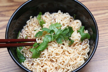 Instant noodles in a black bowl with coriander sprinkles served with chopsticks on a light wooden table.