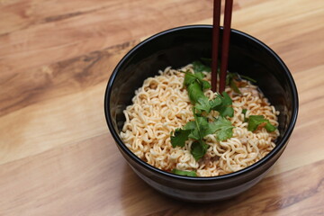 Instant noodles in a black bowl with coriander sprinkles served with chopsticks on a light wooden table.
