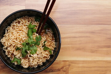 Instant noodles in a black bowl with coriander sprinkles served with chopsticks on a light wooden table.Top view.