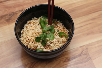 Instant noodles in a black bowl with coriander sprinkles served with chopsticks on a light wooden table.