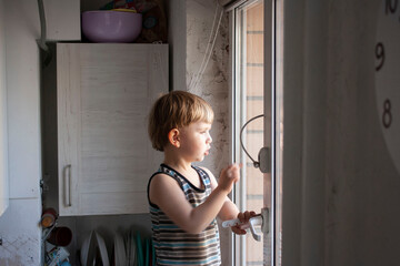 A little boy of 3 years of European appearance stands at the window, on the window there is a protective limiter to prevent falling out of the window
