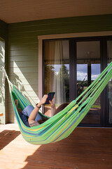 Side view of woman lying in green hammock near cottageand uses a smartphone