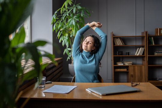 Happy carefree woman stretching hands at workplace smiling relaxed with closed eyes. Millennial student or freelancer journalist writer enjoy break after work done, sitting at desk with closed laptop