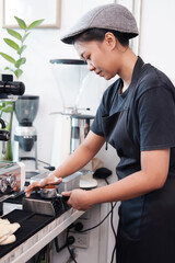 Smiling Asian Young barista woman is wearing apron and brewing coffee with machine for customers order in cafe and coffee shop. Start up small cafe business and technology Concept.