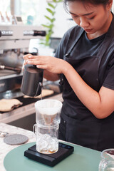 Smiling Asian barista young woman is wearing apron and pouring and craft a hot black coffee into cup for according to the customer's order at counter bar in coffee shop.
