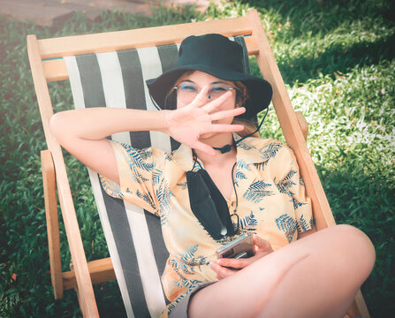 Happy Asian Woman Sitting On A Deck Chair In A Garden With Hot Summer Sunshine.