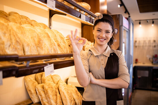 Female bakery worker standing by the shelf full of pastry and bread and holding thumbs up.