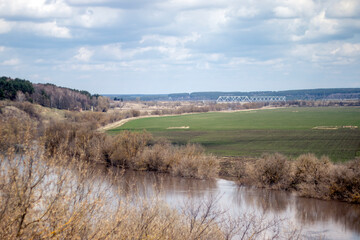 View of the river and fields on a cloudy day in spring. Green grass in the meadows and trees without foliage on the banks of the river. White clouds in the blue sky above the fields.