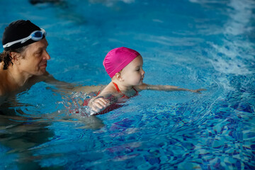 Happy kid toddler learns to swim with coach in the pool, dad grandpa teaches to swim daughter in pink swimming cap