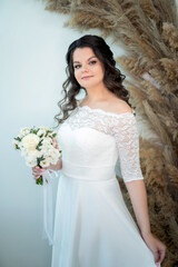 A beautiful bride with curly black hair in a white wedding dress with lace sleeves and a bouquet of white flowers in her hands in the wedding salon