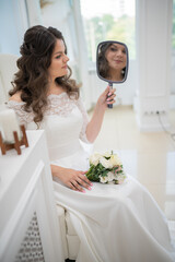 A beautiful bride with curly black hair in a white wedding dress with lace sleeves in the wedding salon is reflected in a mirror that she holds in her hands