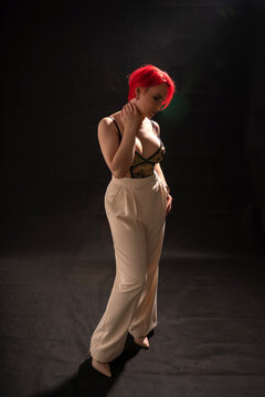 A beautiful woman with large silicone breasts in a lace bra on a bare torso, red short hair, plump lips and light trousers poses for a photographer in a studio standing on a black background