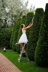 beautiful young woman gardener in a dress cuts pyramidal thujas from a stepladder, topiary art and landscaping in the garden, a classic garden and a girl in a beautiful puffy dress