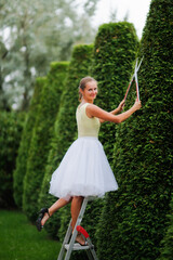 beautiful young woman gardener in a dress cuts pyramidal thujas from a stepladder, topiary art and...