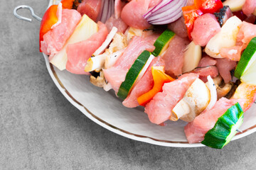 Raw turkey shish kebab on metal skewers, with lots of colorful veggies, on a white plate, ready for preparation. Light grey design desk surface. Delicious, light, great looking food.