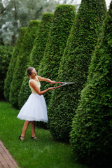 beautiful young woman in a white tulle skirt with garden scissors cuts large pyramidal thuja, garden topiary art. Pretty european woman gardener in a festive dress
