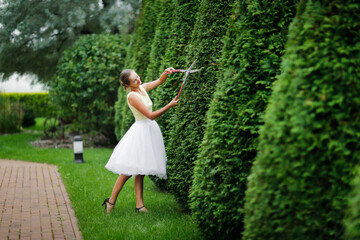 beautiful young woman in a white tulle skirt with garden scissors cuts large pyramidal thuja,...