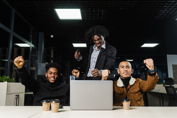 Happy and joyful multiethnic men raise their hands and fists and rejoice in winning in online casino on laptop. Team of black Africans and Asian programmer successful completed project.