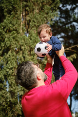 Man having fun with his little son while holding him up in the air with a soccer ball.