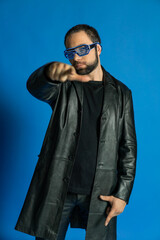 Obraz premium portrait of a man in interactive, cyber glasses. cyberpunk in the studio in black leather clothes. cool trendy handsome guy in flowy leather coat. bearded man with a gun, money on a blue background