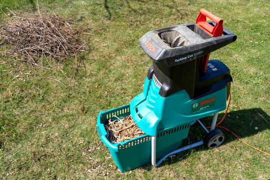 Celadna, Czechia - 04.23.2022: Detail shot of the Bosch electric garden shredder with weathered lawn and pile of cut tree branches in the background. Cleanup around the house. Spring gardening.