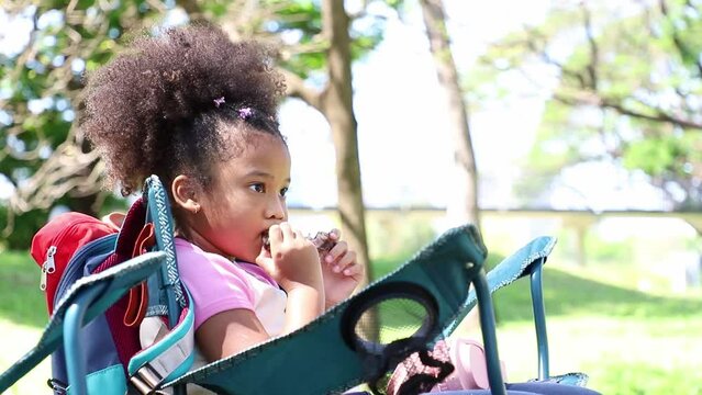Cute Africa American little girl eating grill chicken drumsticks on chair while her camping with family. Ideas for relaxing and eating out
