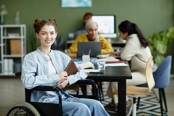 Portrait of smiling young woman in wheelchair looking at camera while working in office with diverse creative team, copy space