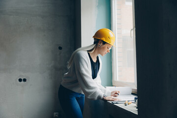 Designer woman in yellow helmet checks author supervision builder work in apartment and compares...