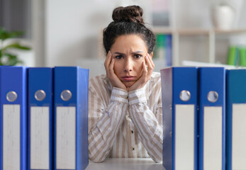 Upset lady businesswoman sitting at workplace with stack of folders and looking at camera, feeling...