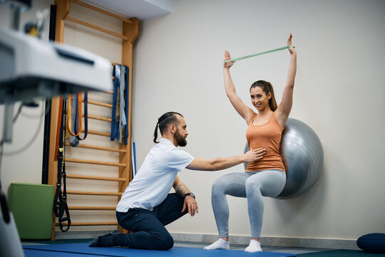Athletic woman uses resistance band and fitness ball while exercising with physical therapist at health club.