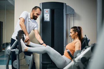 Young sportswoman exercises on leg press machine with help of physiotherapist at health club.
