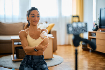 Fitness instructor stretches her arm while recording exercise for her vlog at home.